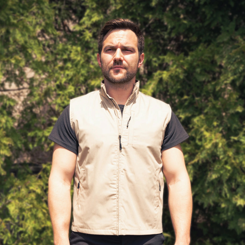 Man wearing a beige vest over a black shirt with green foliage in the background